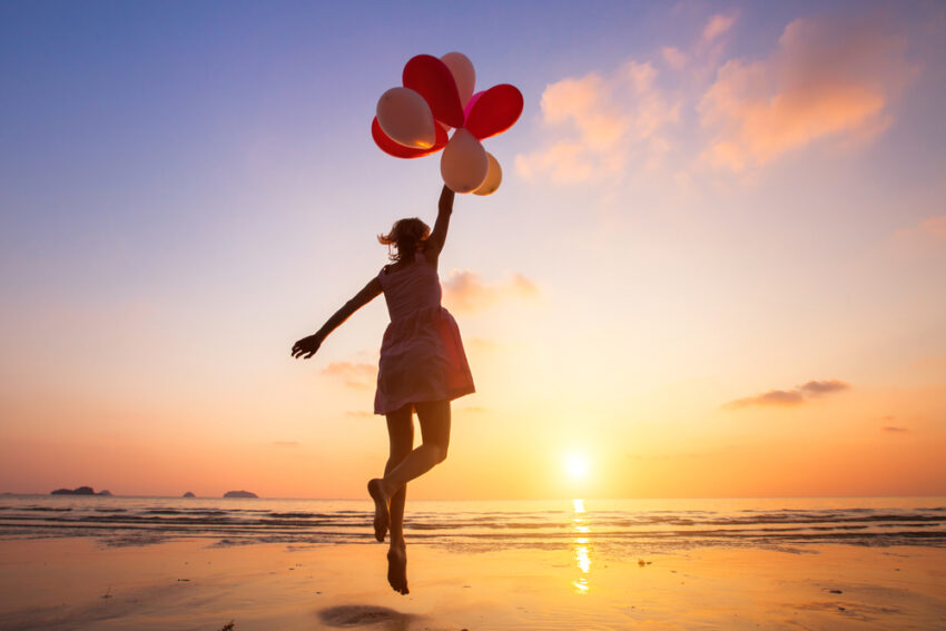 A young girl on a beach floats away with balloons