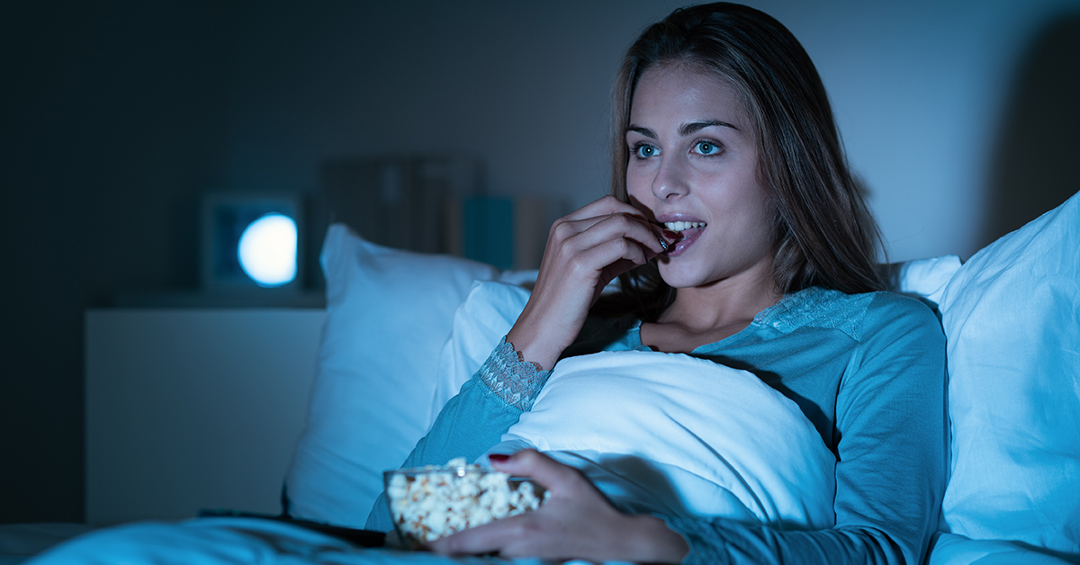 Happy woman relaxing in bed and watching movies on tv late at night, she is eating popcorn